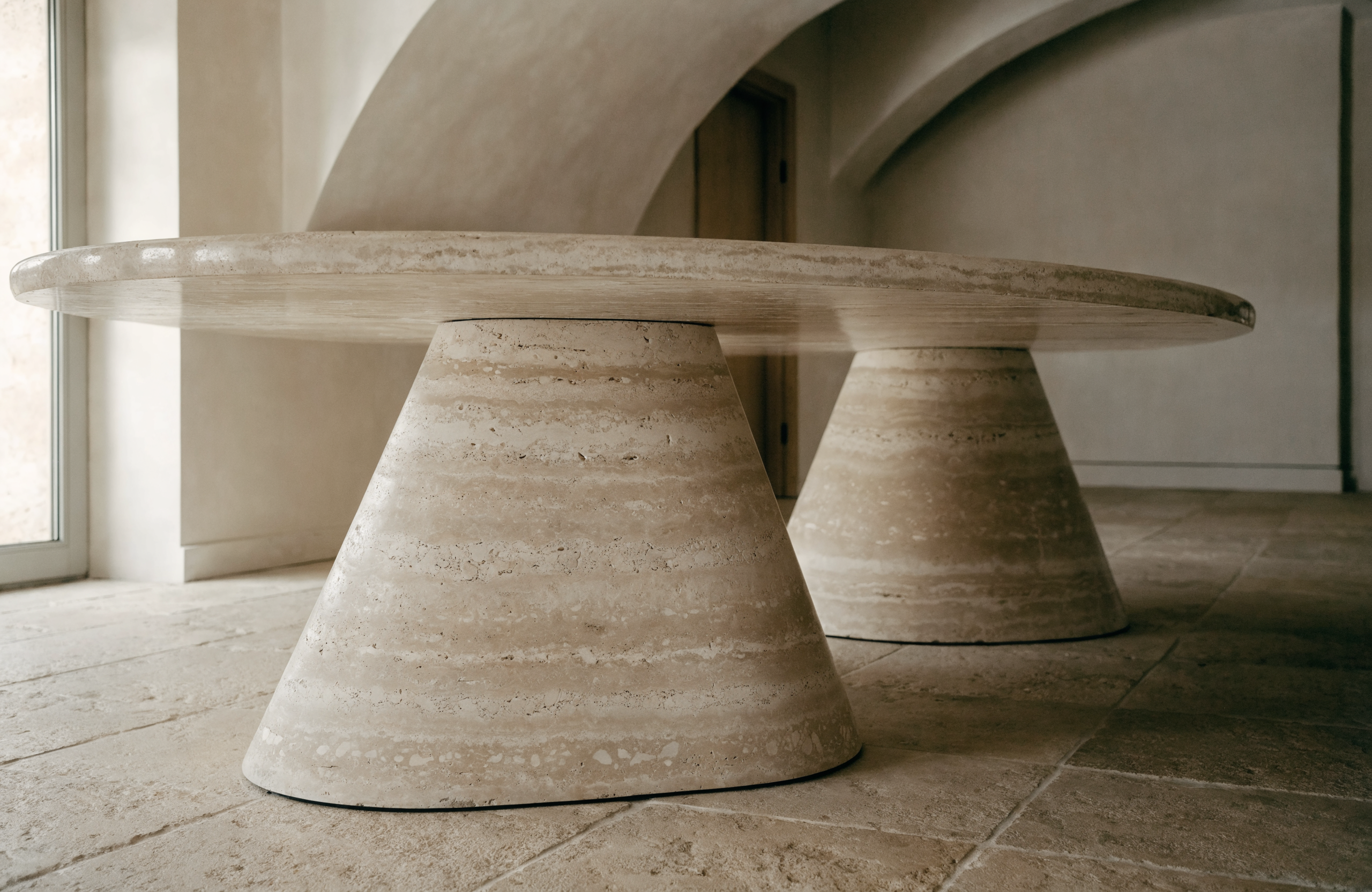 The Oval Travertine Dining Table in dining space — arched plaster ceiling, warm stone floor, Italian editorial